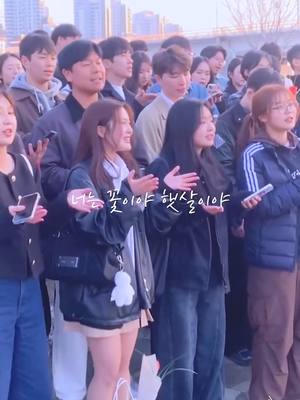 The image features a large group of people, primarily young adults, standing outdoors. Several individuals appear to be singing or gesturing with their hands, suggesting a performance or gathering. The focus is on a cluster of people in the foreground who are actively singing, with other people standing behind them.