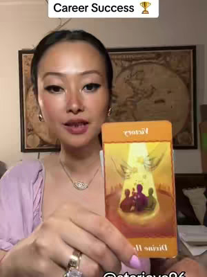 A woman with long dark hair is holding a tarot card. The card depicts an angel figure. The background shows a framed artwork.