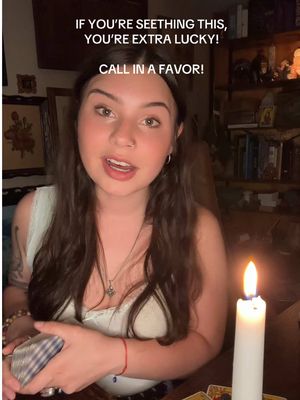A woman with long brown hair and a white top speaks directly to the camera. Text overlays read, "IF YOU'RE SEETHING THIS, YOU'RE EXTRA LUCKY, CALL IN A FAVOR!" A lit candle is to her right and partially visible stacks of tarot cards. Bookshelves and other home items are in the background.