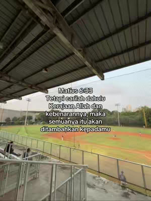 The cover image captures an outdoor setting, possibly a sports field, with a message overlaid. The scene is viewed from a slightly elevated perspective of a stadium or similar structure, framed by a metallic roof and fencing. The field is green with a reddish-brown area. Text is overlaid with biblical verses.