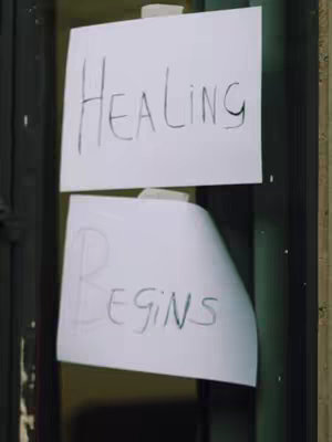 The image shows two pieces of white paper taped to a dark surface. The top paper has the word "Healing" written on it in black marker. The bottom paper has the word "Begins" written on it in black marker.