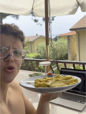 A selfie of a young man holding a plate of food, apparently outside on a bright day. He is sitting at a patio table and looking directly into the camera with a slightly disgusted expression. He's shirtless and visible from the chest up, with glasses. A laptop is open and visible on the table next to him. The background shows trees, houses, and a glimpse of the sea.