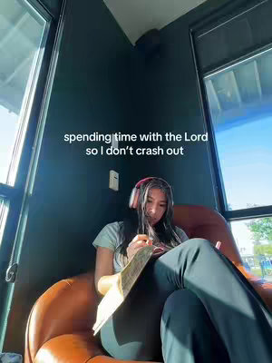 A person sits in a brown armchair indoors, leaning towards the center. The person is wearing a light blue t-shirt, black pants, and light-colored headphones, the person is holding a book open. The background is a dark teal wall with a window in front that has bright sunlight passing through.