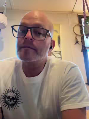 A bald man with a beard and glasses looks at the camera. He is wearing a white t-shirt with a design of a skull with a sun-like pattern around it. He has earbuds in his ears and is in a room with a painting and various hanging plants.