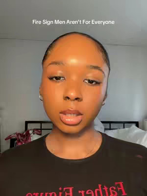 A selfie-style shot features a young woman with dark skin centered and looking directly at the camera. The background is blurred, with a bed frame and bedding faintly visible. She wears a black shirt, and the lighting is even, highlighting her facial features. Text overlays at the top states, 'Fire Sign Men Aren't For Everyone'.