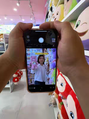 A person is holding a phone displaying a photo of a woman holding a pink teddy bear in a Miniso store. The person holding the phone is in the foreground, their hands framing the phone screen.