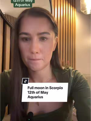 A woman is centered in the frame, looking slightly downward with a neutral expression. The background is a mix of wooden vertical panels with a dim ambient light from the top right. A white rectangle, featuring the text "Full moon in Scorpio 12th of May Aquarius", is overlaid at the bottom middle of the frame. The upper left corner also has text that reads: 12th of May Aquarius. She is wearing a green top and a silver necklace.