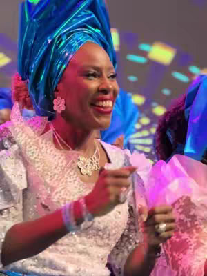 A smiling woman is the focus, dressed in a white and silver textured top and a large, teal-colored headdress. She's wearing large flower-shaped earrings and a sparkly necklace, with bracelets on her wrist, and is pointing her fingers with a joyful expression, with the background slightly out of focus showing a celebration atmosphere in a room.