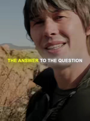 A medium shot features a man with short dark hair and a friendly expression, smiling slightly with teeth visible. He's wearing a dark top that seems to be a jacket, and the background appears to be an outdoor setting, possibly a desert environment with a rocky landscape and a clear sky. Overlaid text in a yellow gradient at the bottom of the frame reads: THE ANSWER TO THE QUESTION. The lighting on the man is bright.