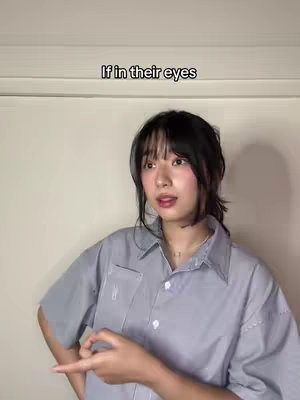 A young woman with dark hair and bangs is shown against a light-colored wall. She is wearing a gray button-down shirt and is gesturing with her right hand, pointing to her left. The video has overlay text at the top.