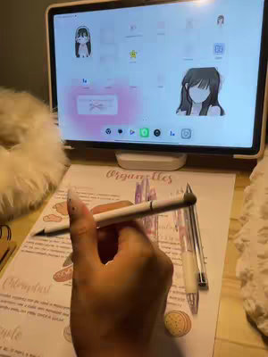 A hand holding a white stylus pen above a sheet of paper with handwritten notes and diagrams. A tablet displaying a digital interface sits in the background. The scene is lit with soft, warm lighting.