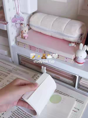 The image shows a desk setup with school supplies. There are school supplies such as books, notebooks, stationary and small decorative objects. The colors in the image are soft and light, giving a calm and aesthetic feel, typically associated with the 'study aesthetic'. A hand is holding a notepad with a page out.