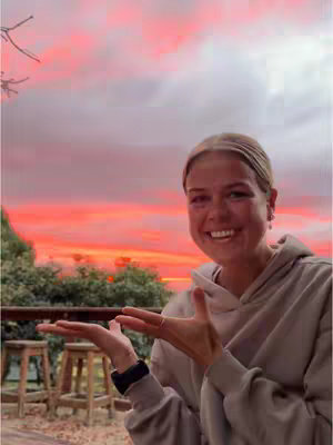 A woman with fair skin and blonde hair is centered in the frame, smiling towards the camera. Her hands are held up, palms facing towards the viewer. The background features a vivid sunset with shades of pink, orange, and white. Behind her is a wooden railing and some greenery, suggesting an outdoor setting. The composition is well-lit, with the sunset providing a warm glow on her face and the scene.
