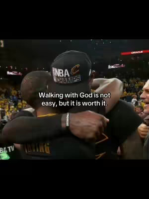 The cover image is a vertical shot from what appears to be a sports arena during a post-game celebration. Two men are embracing. One, with his back to the camera, is wearing a black baseball cap with 'NBA CHAMPS' on it. The other man's face is partially visible, pressed against the first, with a backdrop of cheering people and visible yellow and gold lights. Text overlay at the bottom reads, 'Walking with God is not easy, but it is worth it.' The overall color palette is natural with a slightly desaturated tone, and the composition highlights the emotional connection between the two figures.