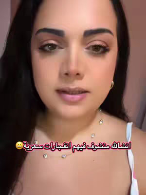 A close-up shot of a woman with dark hair looking towards the camera. She has brown eyes, full lips, and is wearing some makeup, including eyeshadow and mascara. She is also wearing a necklace with several small, clear stones. The background is a soft pink color. The lighting appears to be soft and diffused.