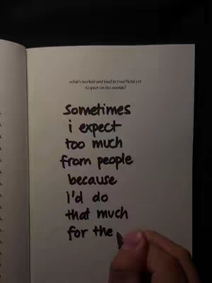 A close-up photo of an open book. On the right page, handwriting states, "Sometimes I expect too much from people because I'd do that much for the..." The photo is taken from a downward angle, with a hand holding a pen positioned at the word "people."