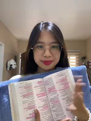 A young woman with long dark hair and glasses smiles while holding open a Bible. The background is a well-lit room with a glimpse of a door and window blinds. She wears red lipstick.