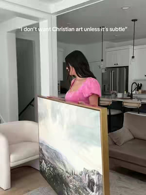 A woman stands in a well-lit room, holding a large framed painting. The painting depicts a landscape, likely a religious scene. The woman is dressed in a bright pink top. The background shows a modern kitchen and living area.