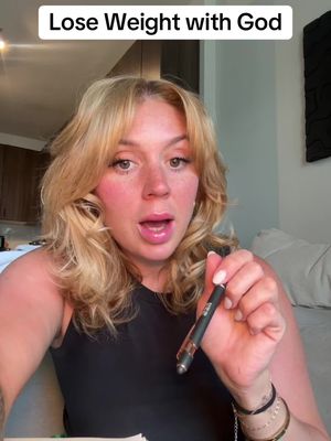 A woman with blonde hair and fair skin is centered in the shot, looking directly at the camera with an open mouth, as if speaking. The upper portion of the image is a text overlay that reads 'Lose Weight with God'. She holds a pen in her right hand and has multiple bracelets on her left wrists. The background is blurred, suggesting an indoor setting with neutral wall colors