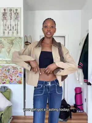 A young woman stands indoors, posing in front of a wall decorated with framed artwork. She wears a black crop top, denim jeans, and a tan jacket with a brown collar. She's pointing down at her stomach, with the text overlayed at the bottom.