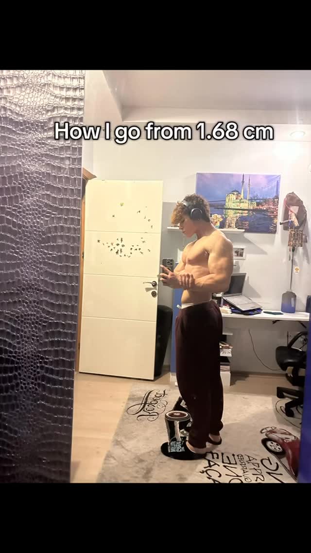 A young man stands shirtless in a room, facing the camera. He is flexing his bicep while holding his phone in one hand. The text overlay shows "How I go from 1.68 cm." The background has a door, a wall with a pattern, a rug, and a desk with a laptop.
