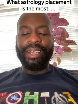 A close-up shot of a Black man with a beard, wearing a dark t-shirt with a colorful logo. He's mid-speech with eyes closed slightly, and the background includes a window with light behind him and some leafy vines.