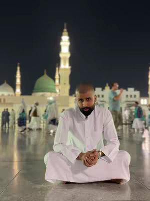 A man in white religious garments sits cross-legged on a stone plaza at night. Behind him, the illuminated architecture of a mosque with tall minarets and a large green dome is visible creating a spiritual ambiance.