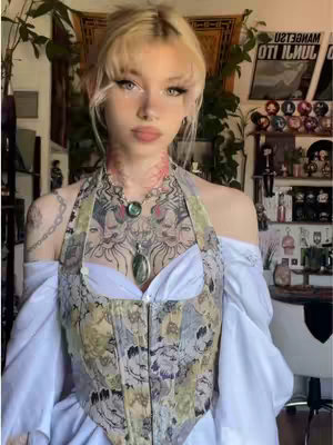 A young woman with blonde bangs wearing a decorative top, jewelry, and off-the-shoulder white sleeves. She is posing in front of a background with plants, a tapestry, and a shelf.