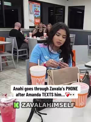 A young woman sits at a table in a fast-food restaurant, looking at her phone. There are two drinks and a purse on the table. Behind her, other people are sitting at tables within the restaurant. The image uses a somewhat saturated color palette, and is moderately lit.
