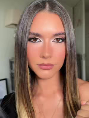 A close-up shot of a woman's face with makeup. She has long, light brown hair with darker roots. Her makeup includes eyeliner, eyeshadow, and pink lip color. The background is softly blurred, showing hints of a room.
