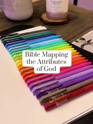 A close-up shot of a collection of colorful gel pens arranged in a rainbow gradient. A white rectangular overlay reads "Bible Mapping the Attributes of God". The pens are laid on a white surface, with the top portion of a wooden table and a white cylindrical object in the background, enhancing the colorful display.