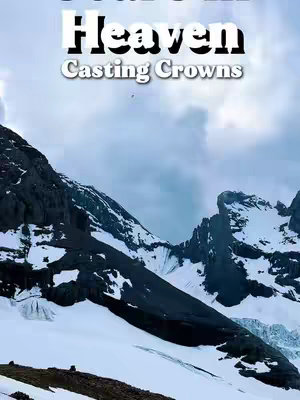 The image is a vertical shot of a mountain scene under a cloudy sky. In the foreground are dark, rocky mountains, with snow patches. In the background stands a tall, snow-covered mountain range with glacial features.  White text overlays are positioned at the top. Overall color palette is comprised of shades of white, gray, and blue.