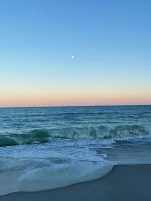 An image of a beach during a sunset or sunrise. The sky transitions from blue at the top to vibrant orange and pink hues near the horizon. The ocean waves are in the foreground, with white foam breaking near the shore. A sliver of the moon is visible in the sky.