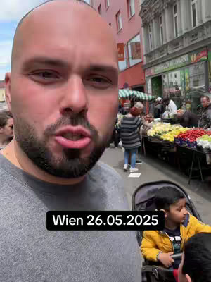 A man with a trimmed beard and shaved head is filming a selfie-style video outdoors. The background shows a street lined with buildings, a market with various products, and other people. A child is visible in a stroller near the man. Text overlaid on the image indicates the location and date.
