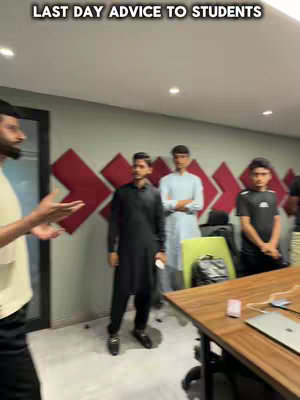 A group of young men are in a room, appearing to be in a mentorship program. The man on the left is speaking, gesturing with his hands. Others are standing. The room has a modern feel with red accents on the wall and a large wooden table in the foreground.
