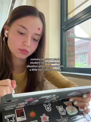 A young woman is shown in a medium shot, seated indoors with a window visible in the background. She holds a tablet computer open in her lap, with stickers visible on the cover. She is wearing a yellow shirt, one earbud in, and gazes downwards with a focused look. A text overlay on the image states, "convinced every psych student has at least one situation when a disorder hits a little too close to home."