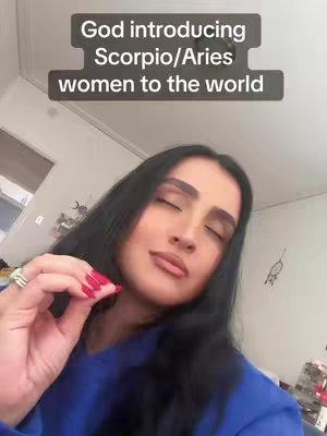 The cover image features a woman with long dark hair, wearing a blue top, in a selfie-style shot. Text overlays read 'God introducing Scorpio/Aries women to the world'. She has her eyes closed and is holding up her left hand, with red nail polish and a ring visible.