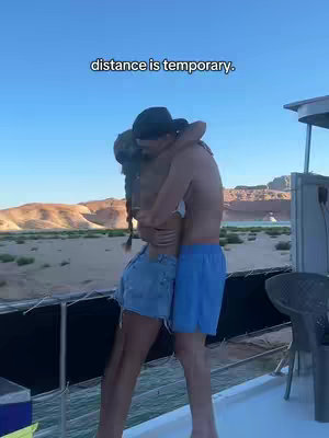 Two people embracing each other on a boat, with a beach and mountains in the background. Overlay text at the top reads 'distance is temporary.'