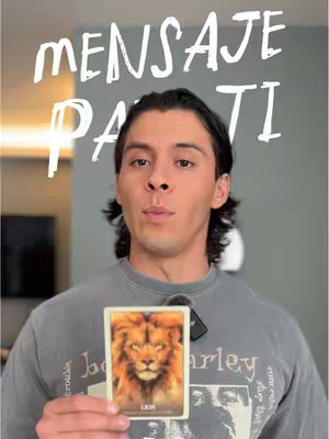 A man with long dark hair is centered in front of a light background. He is wearing a gray graphic t-shirt and holding a tarot card of a lion. Overlayed on the image in stylized white lettering are the words 'MENSAJE PARA TI'.