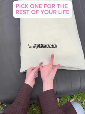 The image shows a first-person perspective of a hand pointing towards a pillow with the superimposed text 'PICK ONE FOR THE REST OF YOUR LIFE' in a rounded rectangle at the top. The hand is positioned above the pillow, which has the number '1. Spiderman' written on it. The background includes a dark surface (likely a couch) and a patch of green grass.