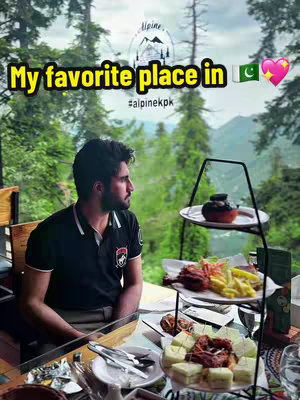 A man sits at a table in an outdoor restaurant with a mountainous backdrop. The man is looking off to the side. A three-tiered serving tray with food is on the table. Text overlaid on the image reads, 'My favorite place in π΅π° π'.