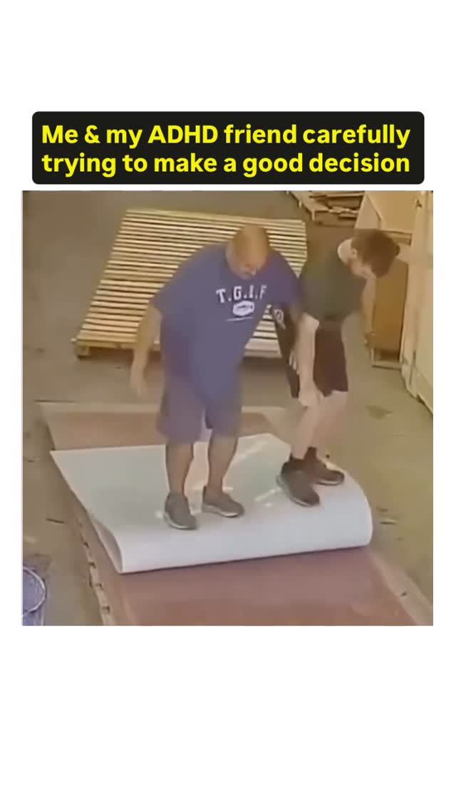 The cover image shows a slightly blurry, medium shot of two men balancing on a large, white, rolled-up surface. They are in what looks like a warehouse or industrial setting. The man on the left, wearing a dark blue t-shirt with white text and dark shorts, is slightly ahead, extending his right arm for balance and looking down. The man on the right, in a dark green shirt and dark shorts, also has his arms out for balance and is looking downward. The background includes wooden pallets and other unseen structures, all bathed in generally dull lighting.