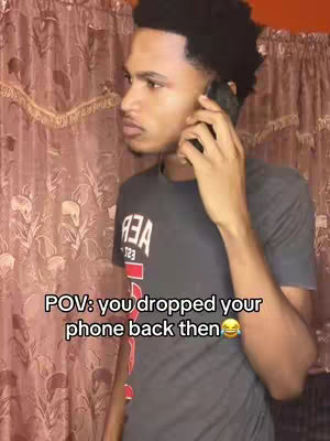 A young man is holding his phone to his ear, looking towards the left side. He has short, dark curly hair and is wearing a gray shirt with white text on it. The background shows a textured, patterned curtain. Text overlay at the bottom reads, "POV: you dropped your phone back then 😂".