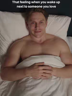 A man with fair skin lies in bed, shirtless and cradling a pillow. He is looking at the camera with a slight smile. The background is dimly lit, hinting at it being morning or evening.