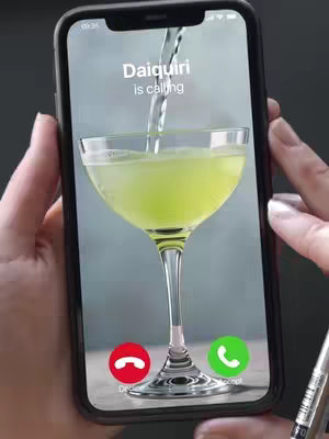 An iPhone is held in the frame, displaying a screen that mimics an incoming call. On the screen, a martini glass is shown, with a stream of liquid pouring into it, presumably to simulate a cocktail being made. The phone screen shows text overlay 'Daiquiri is calling'. Below, red and green buttons, typical of phone call user interfaces, indicate the options to decline or accept the call with the following text prompts: 'Decline' and 'Accept'. The background is a blurry grey. The image is framed by the hands and the phone handle.