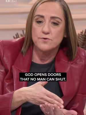 A woman with light brown hair wearing a red jacket speaks in front of a neutral-toned background. Overlayed on the image is a black box containing white text.