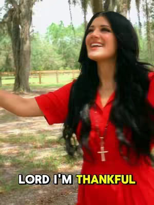 A woman with long black hair and a red dress is standing in a natural setting, possibly a park or a field, smiling and with her arms outstretched. The lower part of the image has a text overlay with the words 'LORD I'M THANKFUL'