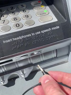 A close-up shot of an ATM keypad with the lower half of the machine visible. A hand is holding a 3.5mm audio jack ready to plug into the machine's port, which is designed for headphones.