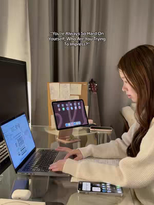 A well-lit, medium shot shows a young woman working at a desk. She's wearing a light-colored sweater and has long, dark hair. In front of her is a laptop open displaying a graph. Beside the laptop is a tablet propped up on a stand. A phone is placed in front of the woman. The background features a curtain and a partially visible desk with a guitar resting against it. Text overlayed above the tablet reads, "You're Always So Hard On Yourself, Who Are You Trying To Impress?"