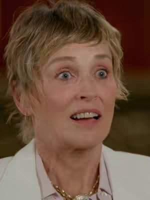 A close-up of Sharon Stone with an expression of surprise. She has short, blonde hair and is wearing a white blazer over a patterned shirt and a gold necklace. Her eyes are wide and mouth slightly open, suggesting shock or amazement. The background is a blurred, warm tone.